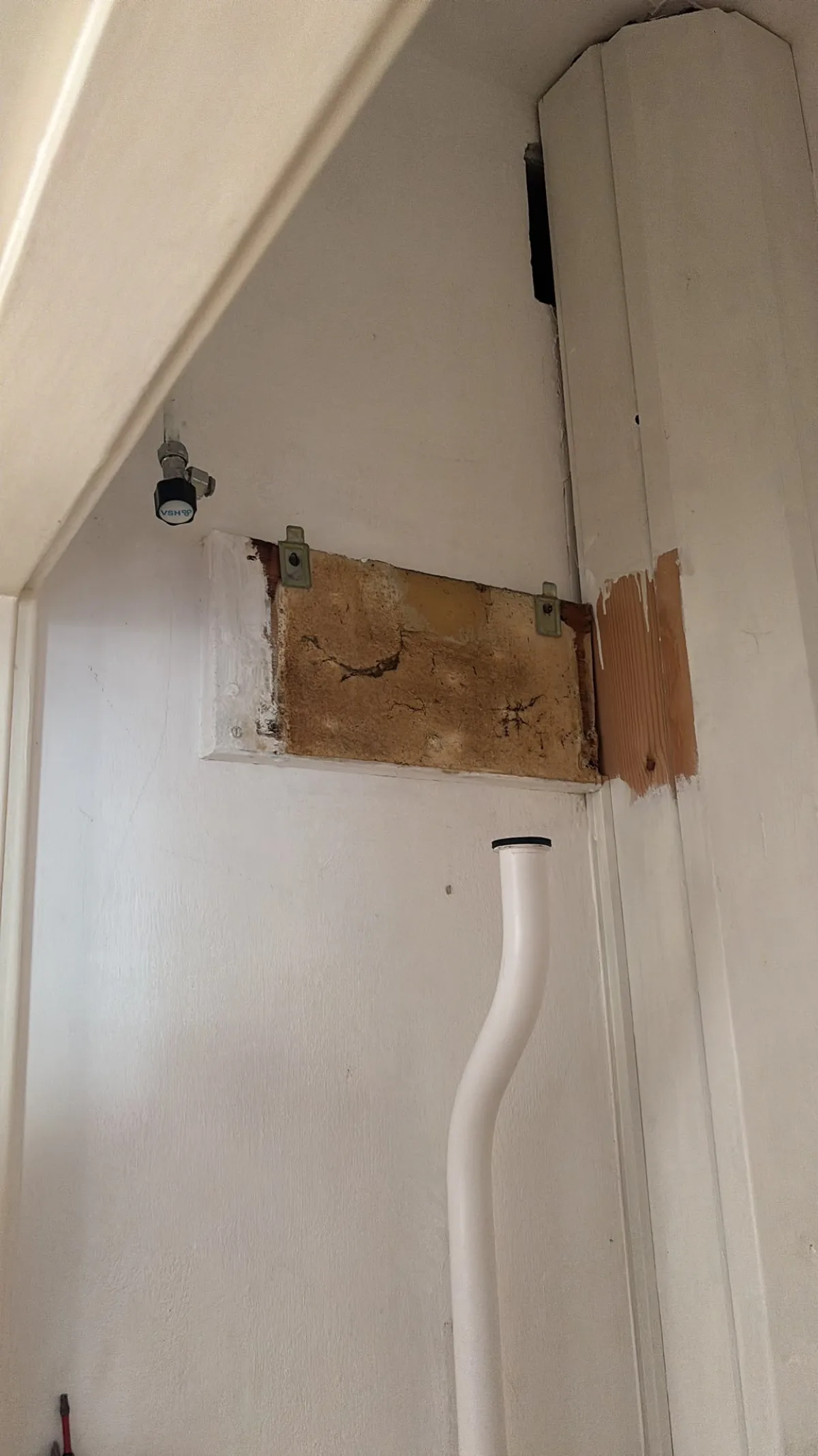 Deels opengewerkte muur met vochtige plekken, blootliggend hout, waterleiding en afvoerbuis.