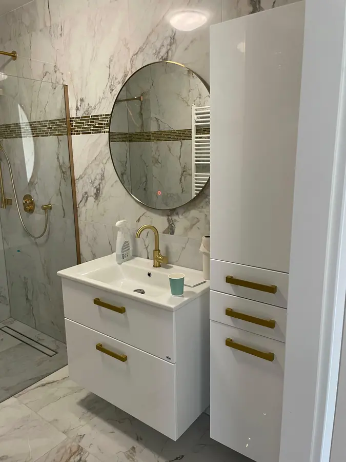 Badkamerrenovatie — Badkamermeubel met wastafel, ronde spiegel en goudkleurige kranen tegen marmerlook tegels